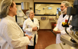 Two women and a man talking. They are all wearing white lab coats and are wearing hairnets