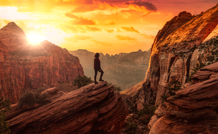 A woman stands on a large rock overlooking a canyon. The rocks and other scenery are full of shades of orange.