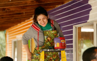 Female volunteer on a ladder with a paint bucket. The volunteer is looking down at someone and smiling.