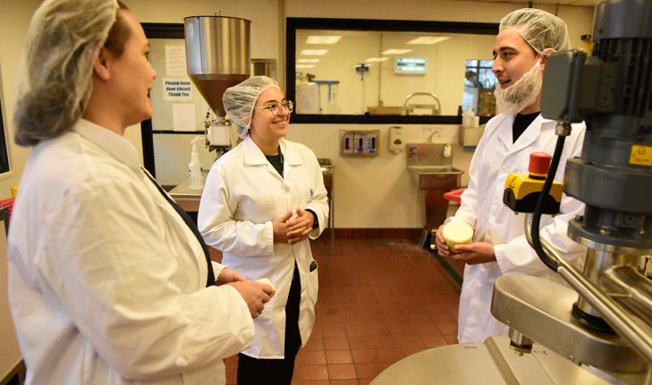 Two women and a man talking. They are all wearing white lab coats and are wearing hairnets