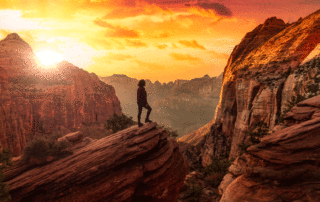 A woman stands on a large rock overlooking a canyon. The rocks and other scenery are full of shades of orange.