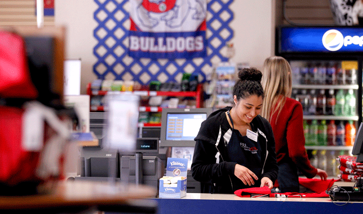 A female Fresno State Bookstore employee standing behind the counter folding shirts.