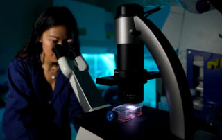 A young woman looks through a microscope in a darkly lit science lab.