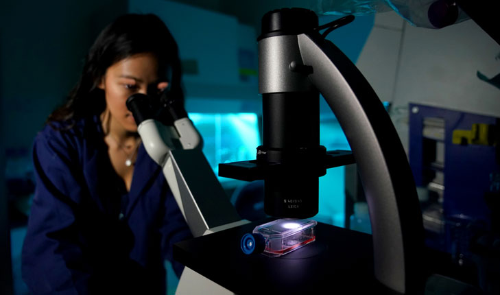A young woman looks through a microscope in a darkly lit science lab.