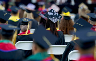 Fresno State graduates sittign with their backs to the camera. A young women's graduation cap is decorated with the words "I did it!"