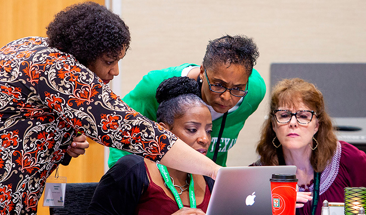 Four women sit around a computer in thought.