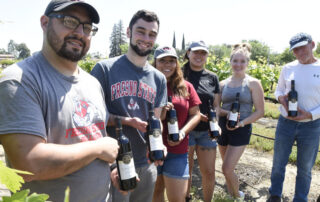 Five Fresno State students pose for a photo with an administrator at the grape fields. They are all holding bottles of Fresno State wine.