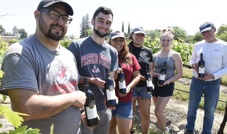 Five Fresno State students pose for a photo with an administrator at the grape fields. They are all holding bottles of Fresno State wine.