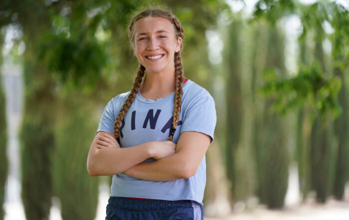 Sydney Fox smiling at the camera. Her hair is in tow long braids and she is wearing a blue shirt.