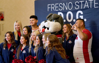 Fresno State cheerleaders and TimeOut stand in front of a Fresno State DOG photo background.