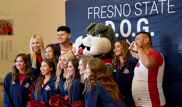 Fresno State cheerleaders and TimeOut stand in front of a Fresno State DOG photo background.