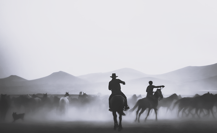 Two riders on horseback guide a herd of horses with mountains rising in the background.