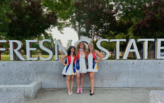 Gladyz Zamora, Caitlin Baten and Amanda Klasen stand together in front of the Fresno State sign with their master's hood and commencement sash.