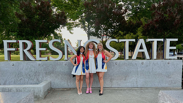 Gladyz Zamora, Caitlin Baten and Amanda Klasen stand together in front of the Fresno State sign with their master's hood and commencement sash.